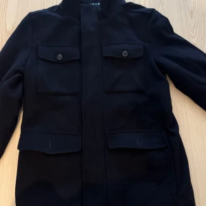 Svart jacka från Dressmann - Svart kappa från Dressmann med fyra fickor framtill och knappar på bröstfickorna. Jackan har lång ärm och är fodrad på insidan för extra värme. Den är endast använd en gång, säljs för 350kr. Nypris ligger på ca. 1700kr 