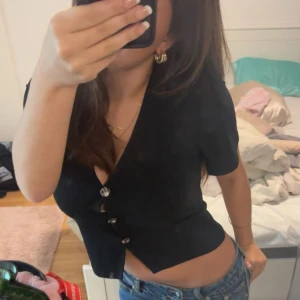 Svart kortärmad blus med knappar - Säljer en svart blus med korta ärmar och snygg v-ringning. Blusen har dekorativa knappar framtill och en figurnära passform som framhäver midjan. Perfekt till jeans eller kjol för en trendig look.