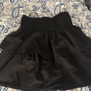 Svart volangkjol med resårmidja - Säljer en svart kortkjol med bred resår i midjan och volangdetalj. Kjolen har en enkel och stilren design som passar perfekt till många olika outfits. Perfekt för dig som gillar en feminin och trendig look.