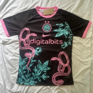Inter Milan fotbollströja Nike x Digitalbits - Säljer en svart Inter Milan fotbollströja med rosa och turkosa detaljer, inklusive ormmönster och växtmotiv. Tröjan har korta ärmar, rosa krage och muddar samt klubbmärke och sponsortryck på bröstet. Finns i S och M