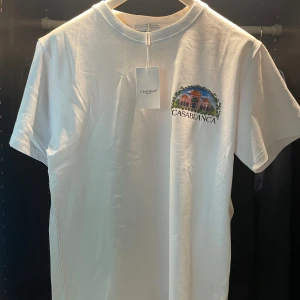 Vit t-shirt från Casablanca med tryck - Vit t-shirt från Casablanca med färgglatt tryck av byggnader och texten 'CASABLANCA' på bröstet och stor illustration på ryggen. Klassisk passform och rund halsringning. Perfekt för dig som gillar stilrena men ändå unika plagg.