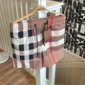 Rutig badshorts från Burberry - Snygga badshorts från Burberry med klassiskt rutigt mönster i beige, svart, vit och röd. Shortsen har snörning i midjan och är tillverkade i ett lätt syntetmaterial. Perfekta för sommaren och strandhäng.