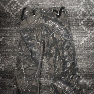 Cargobyxor Camoflage Baggy XXL - Säljer ett par cargobyxor med grått och svart camouflage-mönster. Byxorna har flera fickor, justerbara spännen i midjan och raka ben. Perfekta för dig som gillar en tuff stil och vill ha praktiska byxor med mycket förvaring.