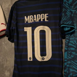Frankrike Mbappé fotbollströja - Säljer en blå och svart randig Frankrike fotbollströja med Mbappé och nummer 10 på ryggen. Tröjan är kortärmad och har klassiska blå nyanser med tunna svarta ränder. Perfekt för fans av landslaget eller Mbappé.