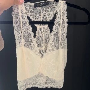 Vit spetsig croppad linnetopp från Soaked - Säljer en superfin vit croppad linnetopp från Soaked i spets. Toppen är ärmlös med v-ringning och vackra spetsdetaljer över hela plagget. Perfekt att styla till jeans eller kjol för en trendig look.