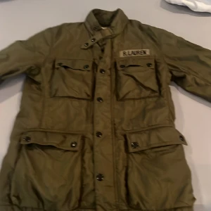 Olivgrön jacka från Denim & Supply Ralph Lauren - Säljer en olivgrön jacka från Denim & Supply Ralph Lauren. Jackan har fyra fickor framtill med lock och knappar, hög krage med spänne och knappstängning. Klassisk militärinspirerad look med patch på bröstet.
