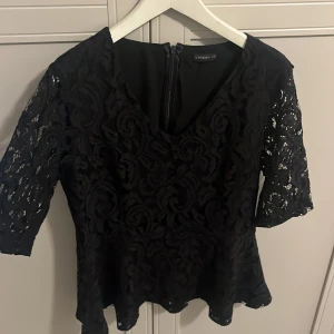 Svart spetsblus från Stockholm  - Säljer en svart blus från Stockholm med vackra spetsdetaljer och halvärmade ärmar. Blusen har v-ringning och dragkedja i ryggen. Perfekt för dig som vill ha en elegant och stilren topp med fina mönster.