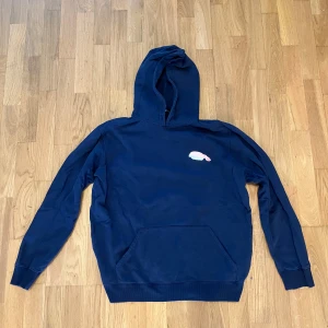 Mörkblå hoodie från Axel Arigato med sushibroderi - Säljer en mörkblå hoodie från Axel Arigato med ett broderat sushimotiv på bröstet. Tröjan har huva och en stor magficka framtill. Perfekt för dig som gillar streetwear och unika detaljer. Storlek L