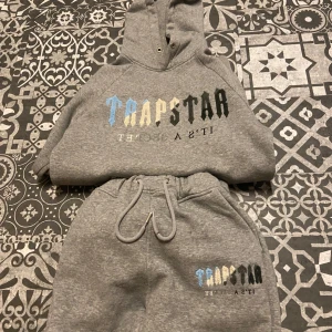 Grå hoodie och mjukisbyxor från Trapstar - Säljer ett grått set från Trapstar med hoodie och matchande mjukisbyxor. Båda delarna har färgglad Trapstar-logga i blått, gult och svart framtill. Hoodien har dragsko i huvan och byxorna har resår och dragsko i midjan.