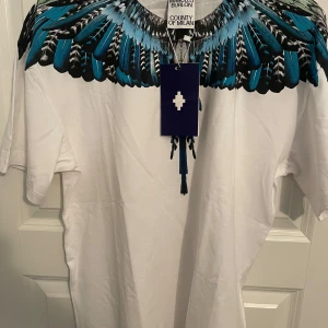 Vit t-shirt Marcello Burlon med fjädertryck - Snygg vit t-shirt från Marcelo Burlon County of Milan med ikoniskt blått och svart fjädermönster runt halsen. Klassisk passform och korta ärmar, perfekt för dig som vill sticka ut med en cool design. Materialet är mjuk bomull.