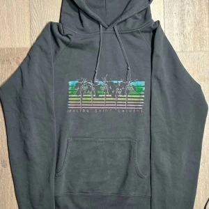 Mörkgrå hoodie med tryck från Malibu Saint Laurent - Säljer en mörkgrå hoodie med tryck av palmer och gröna detaljer från Malibu Saint Laurent. Tröjan har huva med dragsko och en stor magficka. Perfekt för en avslappnad stil.