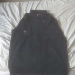 Svart pikétröja från Lacoste - Säljer en klassisk svart pikétröja från Lacoste med broderad krokodillogga på bröstet. Tröjan har korta ärmar, krage och knappar framtill. Perfekt för en stilren och sportig look.