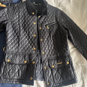 Svart quiltad jacka från Barbour - Säljer en svart quiltad jacka från Barbour med guldiga knappar och dragkedja. Jackan har klassisk krage i manchester, flera fickor framtill och Barbour-logga på ena fickan. Perfekt för våren😊😍 passar mig bra och har S/M och är 163cm lång🥰