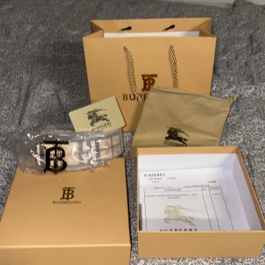 Vändbart bälte från Burberry med klassiskt mönster - Säljer ett vändbart Burberry-bälte med stor metallspänne i svart. Ena sidan har Burberrys rutiga mönster i beige, grått och vinrött, andra sidan är svart med diskret rutmönster och ingraverad logga. Kommer med originalförpackning och dustbag.