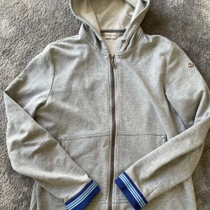 Grå Moncler tröja  - Säljer en grå Moncler tröja med dragkedja och blå-vita ribbade muddar vid ärmsluten. Tröjan har huva och fickor framtill samt Moncler-logga på ärmen. Skick 6,5/10 size L fits S/M