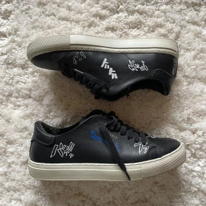 Svarta sneakers från Axel Arigato - Säljer ett par svarta sneakers från Axel Arigato med vita och blå grafiska tryck på sidorna. Lite slitningar (bild 5) annars är de knappt använda. Storlek 38. Skriv för frågor!☺️