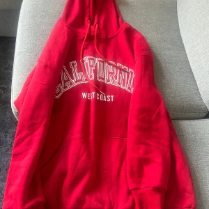 Röd hoodie med tryck 'California West Coast' - Säljer en röd hoodie med vit text 'California West Coast' på bröstet. Tröjan har huva med dragsko och en stor magficka framtill. Perfekt för en avslappnad stil.