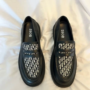 Dior Explorer loafers - Säljer ett par svarta loafers från Dior med kraftig sula och mönstrad textil på ovandelen. Skorna har rund tå och platt sula, samt klassisk penny loafer-detalj framtill. Perfekta för dig som vill ha en stilren men unik look.