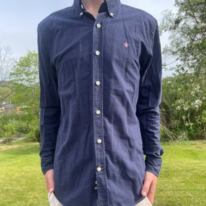 Mörkblå skjorta från Morris - Säljer en klassisk mörkblå skjorta från Morris i storlek M. Skjortan har knappar hela vägen, button-down krage och en liten broderad logga på bröstet. Perfekt för dig som gillar stilrena och tidlösa plagg. Den har ett pyttelitet hål i nacken som knappast syns!