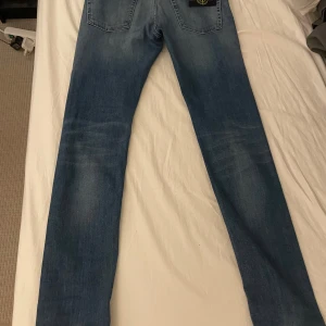 Blå jeans från Stone Island Junior - Säljer ett par klassiska blå jeans från Stone Island Junior med raka ben och snygg tvätt. Byxorna har bakfickor och den ikoniska Stone Island-loggan på ena bakfickan. Perfekta för dig som gillar stilrena jeans med en exklusiv touch.
