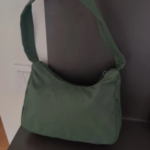 Grön axelväska från Gina Tricot - Snygg grön axelväska från Gina Tricot med enkel design och bred axelrem. Väskan har dragkedja upptill och är tillverkad i ett mjukt syntetmaterial. 