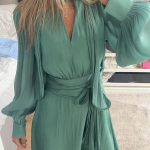 Grön omlottklänning med knytband - Snygg grön klänning med omlottkänsla och brett knytband i midjan. Klänningen har långa, vida ärmar med resår vid handlederna och v-ringad hals. Perfekt för dig som gillar en flowy och elegant stil. Aldrig använd! 