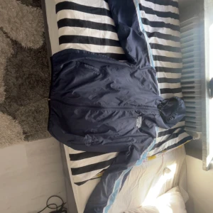 Blå vindjacka med Norra läktaren Malmö-tryck - Säljer en blå vindjacka med huva och dragkedja framtill. Jackan har ett stort tryck på bröstet med texten 'Norra läktaren Malmö' och ljusblå detaljer längs ärmarna. Perfekt för dig som vill visa ditt stöd på läktaren.