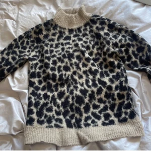 Leopardmönstrad stickad polotröja - Säljer en stickad polotröja med leopardmönster i svart och beige. Tröjan har hög krage och långa ärmar samt ribbade muddar vid hals, ärmslut och nederkant. Perfekt för dig som vill sticka ut lite extra!