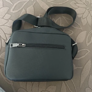 Svart axelväska från Lacoste - Snygg svart axelväska från Lacoste med justerbart band och silverfärgad dragkedja. Väskan har en ytterficka med dragkedja och en diskret Lacoste-logga framtill. Perfekt för att bära dina viktigaste saker.