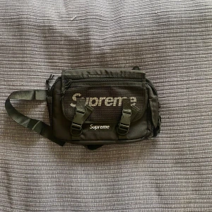 Svart axelväska från Supreme - Säljer en svart slingbag från Supreme med justerbar rem och flera fack. Väskan har två spännen framtill, dragkedja och Supreme-logga på framsidan. Perfekt för att bära med sig lätta mängder med saker, har även plats för vissa större saker. Priset ej hugget i sten. Aldrig använd.