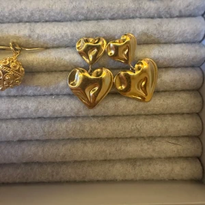 Ioaku örhängen  - Ett par glansiga örhängen i guldton, formade som hjärtan med en mjukt böljande design. Perfekta för att addera lite extra kärlek till din look. Aldrig använda