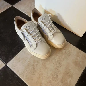 Guiseppe Zanotti skor - Säljer ett par vita sneakers från Giuseppe Zanotti med snörning och dragkedjor på sidorna. Skorna har rund tå, platt sula och är tillverkade i skinn. Perfekta för dig som gillar stilrena och exklusiva sneakers.