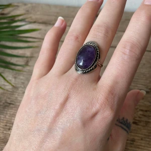 Silverring med ametist - Säljer en snygg silverring med en stor oval lila  ametist. Ringen har en dekorativ tvinnad kant runt stenen och fina detaljer på sidorna. Perfekt statement-piece för dig som gillar unika smycken. Ställbar