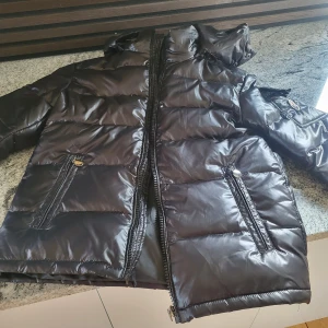 Svart pufferjacka med huva - Säljer en svart pufferjacka med huva och dragkedja. Jackan har två fickor framtill med dragkedja och knappdetalj. Perfekt för kyligare dagar och riktigt skön att bära.