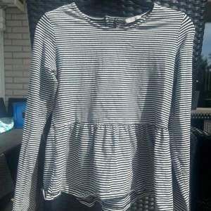 Randig långärmad topp från Lindex Kids - Säljer en svartvit randig långärmad topp från Lindex Kids. Toppen har rund halsringning, dragkedja i ryggen och en lätt volang nedtill. Perfekt för dig som gillar enkel men snygg stil.