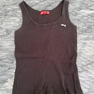 Svart ribbad linnetopp från Puma - Säljer en svart ribbad linnetopp från Puma med klassisk logga broderad i vitt på bröstet. Toppen har en rundad halsringning och är ärmlös, perfekt för en sportig och avslappnad stil.