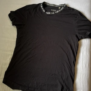 Svart t-shirt från Guess med logga i kragen - Svart t-shirt från  Guess med vitt märke runt kragen.Tröjan är i storlek S och har en plain look.