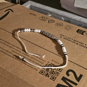 Vitt armband med pärlor och mönstrad detalj - Säljer ett vitt armband med små pärlor och en större mönstrad pärla i mitten. Armbandet har justerbar knytning och inslag av gråa detaljer. Perfekt accessoar för en stilren look.