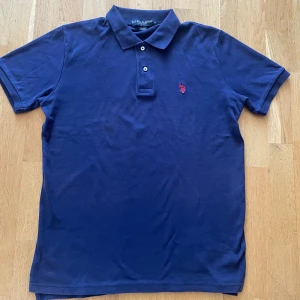 Marinblå pikétröja från U.S. Polo Assn. - Tjena! Säljer en klassisk marinblå pikétröja från U.S. Polo Assn med röd broderad logga på bröstet. Tröjan har korta ärmar, krage och två knappar framtill. Perfekt för en stilren och avslappnad look. Tröjan är knappt använd och är i väldigt bra skick. Hör av dig vid minsta fundering! 