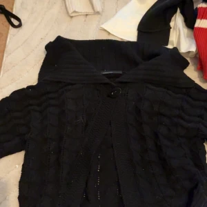 Svart stickad kofta med krage - Säljer en svart stickad kofta med korta ärmar och stor krage. Koftan har ett snyggt strukturmönster och stängs med en knapp framtill. Perfekt att slänga över en topp för en cool look.