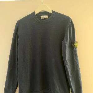 Mörkblå tröja från Stone Island - Säljer en mörkblå långärmad tröja från Stone Island med klassisk rund halsringning och den ikoniska logotypen på vänster ärm. Tröjan har en enkel och stilren design, perfekt för dig som gillar streetwear och snygga basplagg.