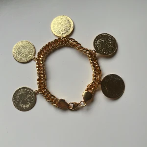 Guldarmband med myntberlocker - Snyggt armband i guld-look med fem stora myntberlocker. Kedjan är 21 cm lång och 2,5 cm bred. Perfekt accessoar för att ge din outfit en unik touch. Aldrig använd. 