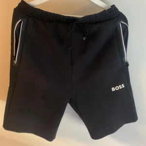Svarta shorts från BOSS  - Svarta shorts från BOSS med vita dragkedjor på fickorna och snörning i midjan. Klassisk design med logga på ena benet. Perfekta för en avslappnad stil.
