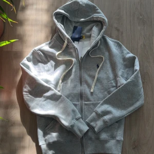 Grå hoodie från Polo Ralph Lauren - Snygg grå hoodie från Polo Ralph Lauren med dragkedja och vit dragsko. Tröjan har broderad logga på bröstet och är tillverkad i mjuk bomullsmix. Perfekt för en avslappnad stil och enkel att matcha med andra plagg.