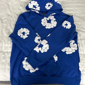 Blå hoodie med vita blommor från Denim Tears - Säljer en blå hoodie från Denim Tears med vita blommönster över hela tröjan. Tröjan har huva och en klassisk känguruficka framtill. Perfekt för dig som vill sticka ut med en unik och färgglad stil. Storlek L men passar även till M