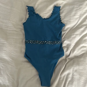 Blå baddräkt med volangkant och leoparddetalj - Använd 1 gång men absolut inga skador eller sånt. Storlek 176 så som xs. Säker pågrund av för litet för mig.Säljer en blå baddräkt med volangkant längs axelbanden och ett leopardmönstrat band i midjan. Materialet är ribbat och baddräkten har en klassisk passform. Perfekt för strand eller pool!