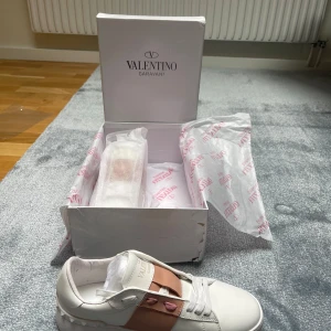 Valentino Garavani sneakers med rosa detalj - Säljer ett par snygga vita Valentino Garavani sneakers med bred rosa rem och nitar på sidan. Skorna har rund tå, platt sula och klassisk snörning. Perfekta för dig som vill ha en stilren men ändå unik look.