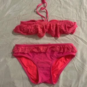 Rosa bikini med volangdetaljer - Söt rosa bikini med volang och spetsdetaljer på både överdel och underdel. Överdelen har knyt i nacken och spänne i ryggen. Materialet är syntet och färgen är starkt rosa. Perfekt för strand eller poolhäng! Lite solblekt.