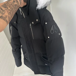 Svart dunjacka med pälskant från Moose Knuckles - Säljer en svart dunjacka från Moose Knuckles med silverfärgade dragkedjor och pälskant på huvan. Jackan har flera fickor med dragkedja och tryckknappar samt logga på ärmen. Perfekt för kalla dagar. Stor på mig och är 174 