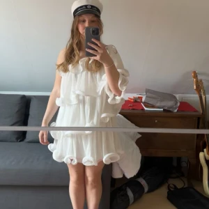 Vit volangklänning med oneshoulder - Superfin vit kortklänning med volanger och oneshoulder-design. Aldrig använd, med prislapp kvar. Säljer då jag har annan klänning till studenten!  Klänningen har en luftig och flowy passform markerade volangdetaljer längs hela klänningen. Perfekt för student eller till sommaren💕